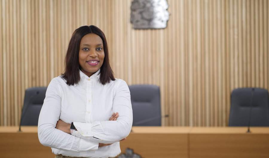 A person stands in a courtroom with their arms folded. In the background is a bench with a crown seal above it.