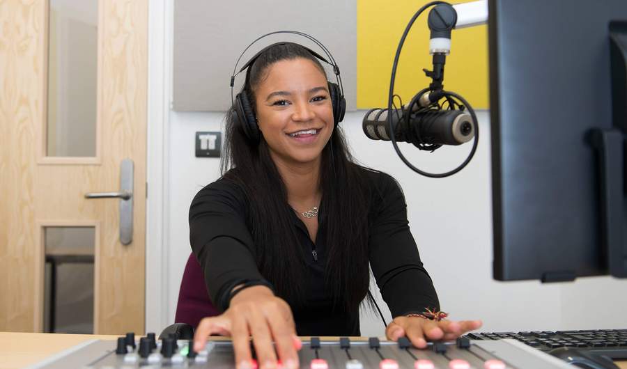 A person wearing headphones sits at a sound mixing desk. In front of them is a microphone mounted on an adjustable arm.