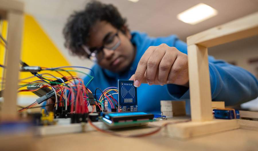 A person in a blue hoodie works with electronic components and circuit boards, placing a small blue board among connecting wires on a table. The background is blurred and the lighting is bright.