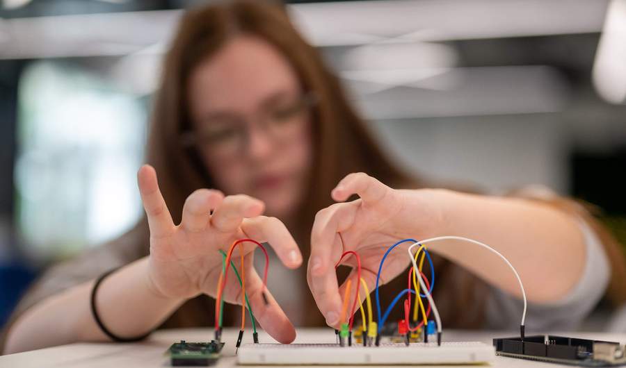 A person with long hair and glasses is working with a breadboard and wires, connecting electronic components. The background is blurred, focusing on the hands and the breadboard.