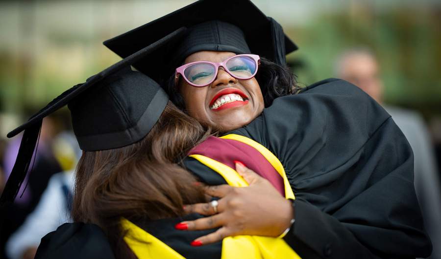 Two students in graduation caps and gowns hug each other.