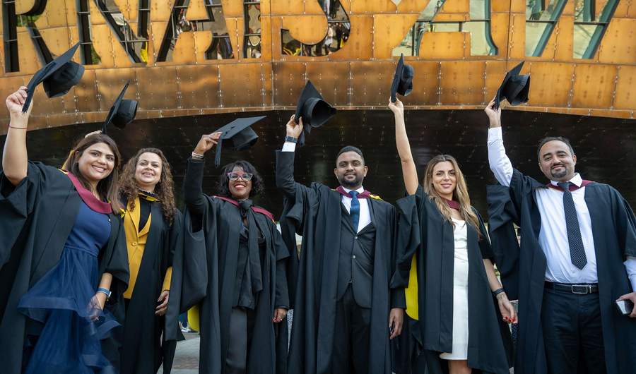 Six students in graduation gowns line up in front of the Wales Millennium Centre, holding their caps in the air.