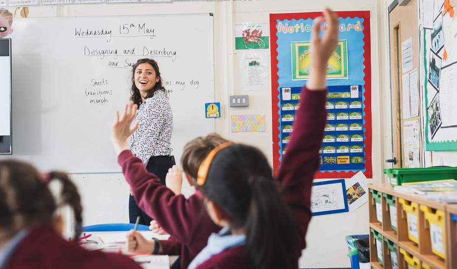 A teacher writes on a whiteboard while two pupils raise their hands.
