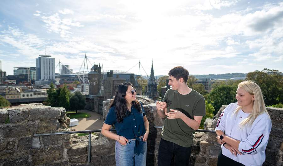 Three people converse while standing on the wall of a castle, with a panoramic view of a city in the background.