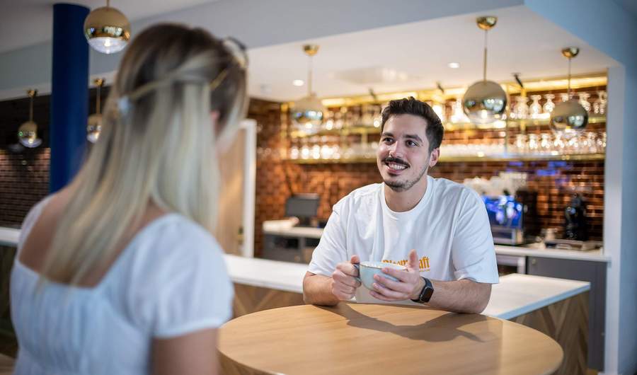 Two people sit at a table talking to each other. In the background is a bar area with a coffee machine.