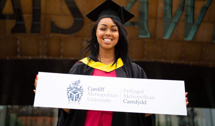 A Cardiff Met student, in graduation cap and gown, stands holding a sign featuring the Cardiff Met logo and name