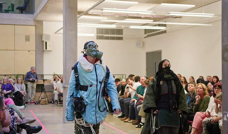 Two models walk along a fashion catwalk. They are wearing heavy coats and gas masks.