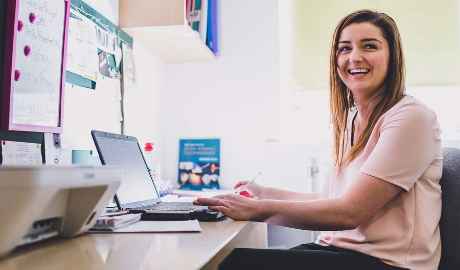 Smiling woman working at her desk with a laptop, taking notes in a bright office space.