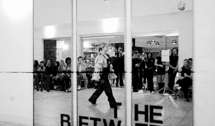 A black and white image of a mirror in which the reflection of a catwalk fashion show with attendees can be seen