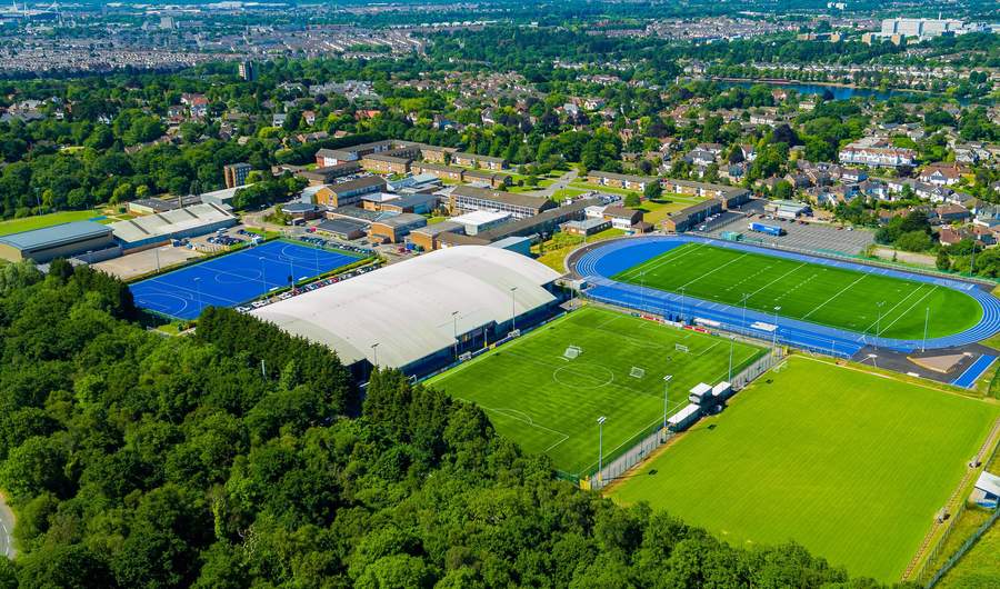 Aerial view of a sports complex featuring a large indoor sports facility, blue athletics track, multiple soccer fields, lush green trees in the foreground, and nearby residential buildings in the background.