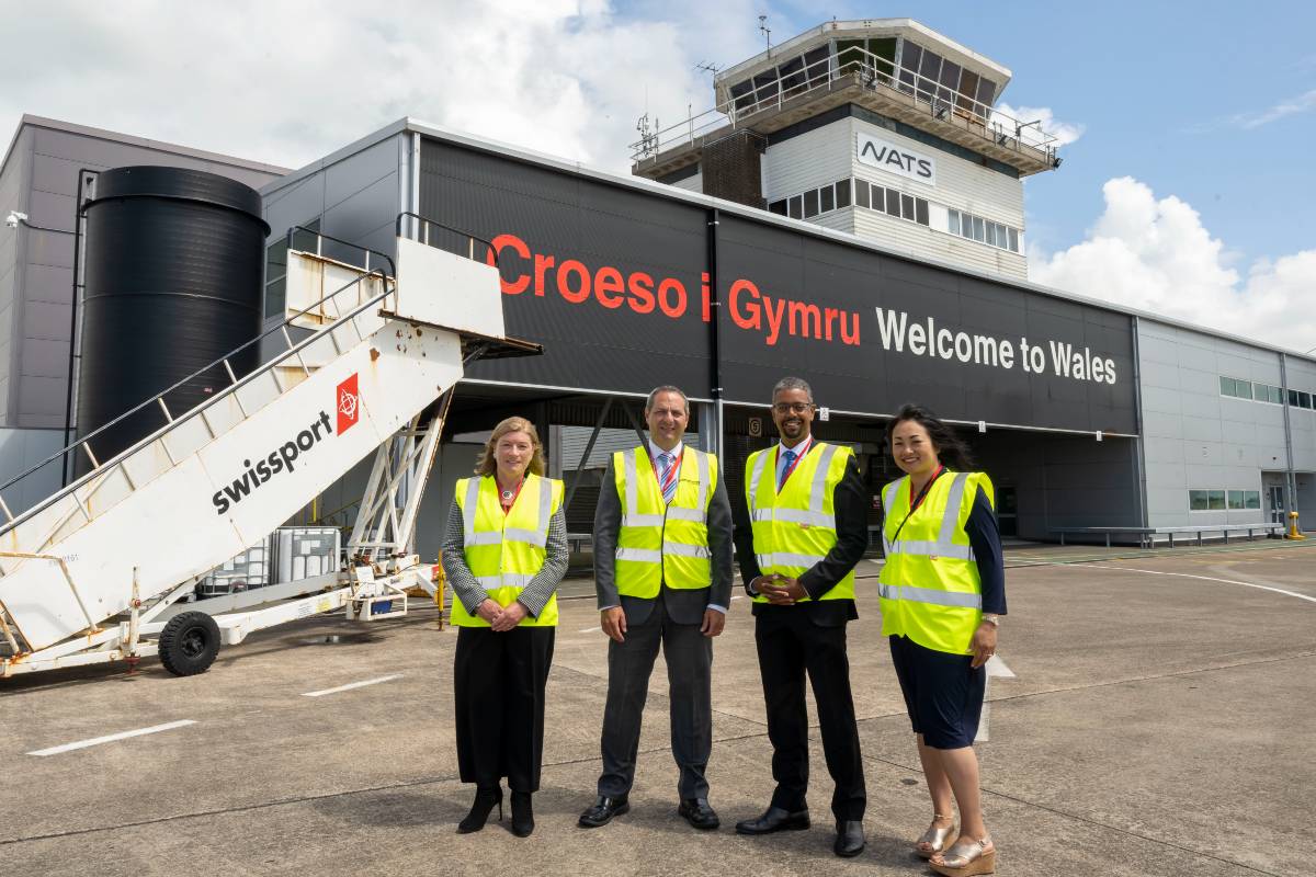 Cara Aitchison, Spencer Birns, Vaughn Gething and Nancy Tran-Horne wearing high-visibility jackets in front of the Cardiff Airport welcome sign