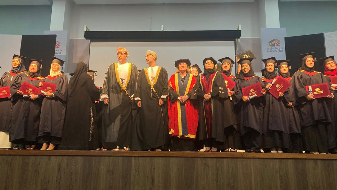 Gulf College Oman graduates stand on stage in their graduation gowns and caps holding their degrees