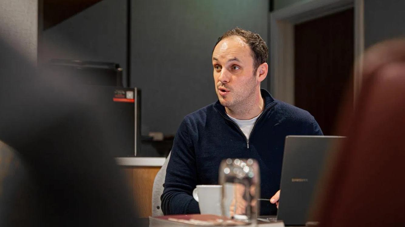 Tomos Lewis speaking during a meeting, seated at a table with a laptop and coffee cup in an office setting.
