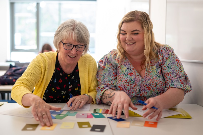 Two women work at a desk sorting coloured cards