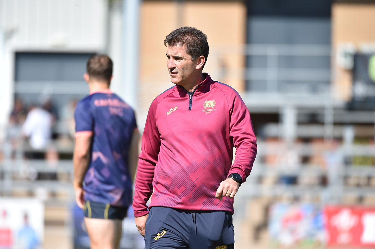 A man wearing a red shirt and blue shorts strolls on a green field, coaching rugby players.