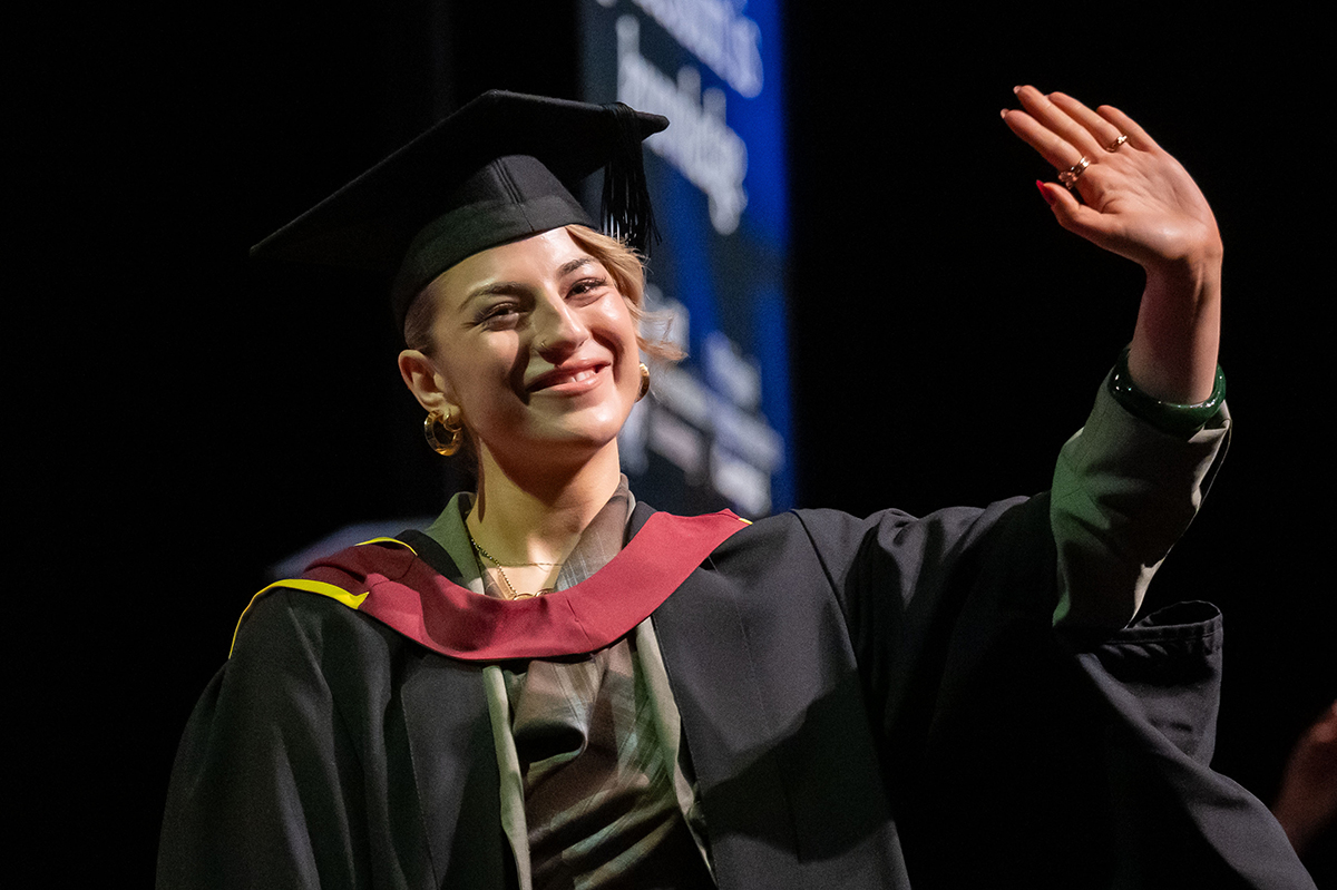 A woman in a graduation gown waves joyfully at the audience during her graduation ceremony.