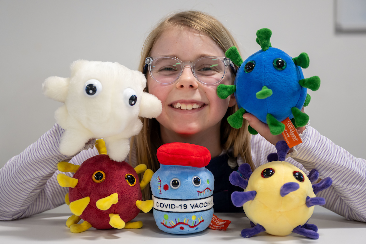 A young girl wearing glasses poses for a photograph surrounded by a variety of colourful plushy toys