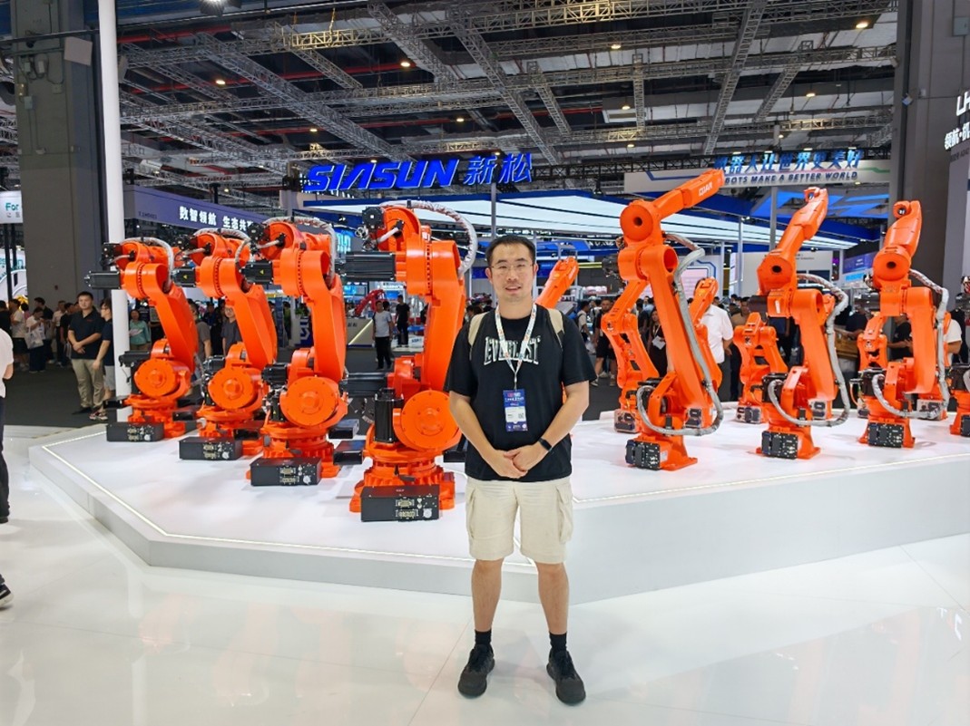 At an exhibition, a man poses in front of several orange robots, highlighting advancements in robotics and automation.