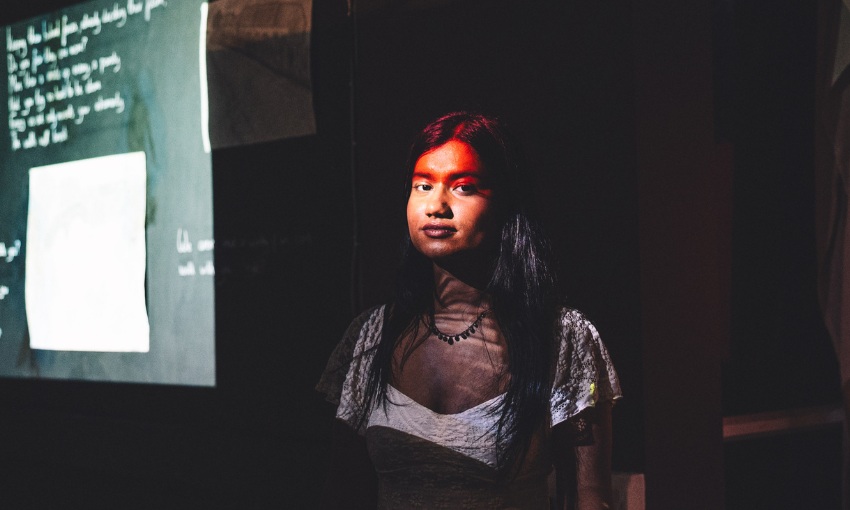 A young woman looks at the camera in a low light room, illuminated by the light from a projector