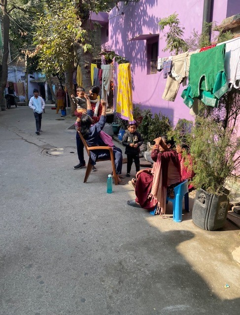 People in traditional Indian attire sit on the sidewalk beside a purple building and a tent.