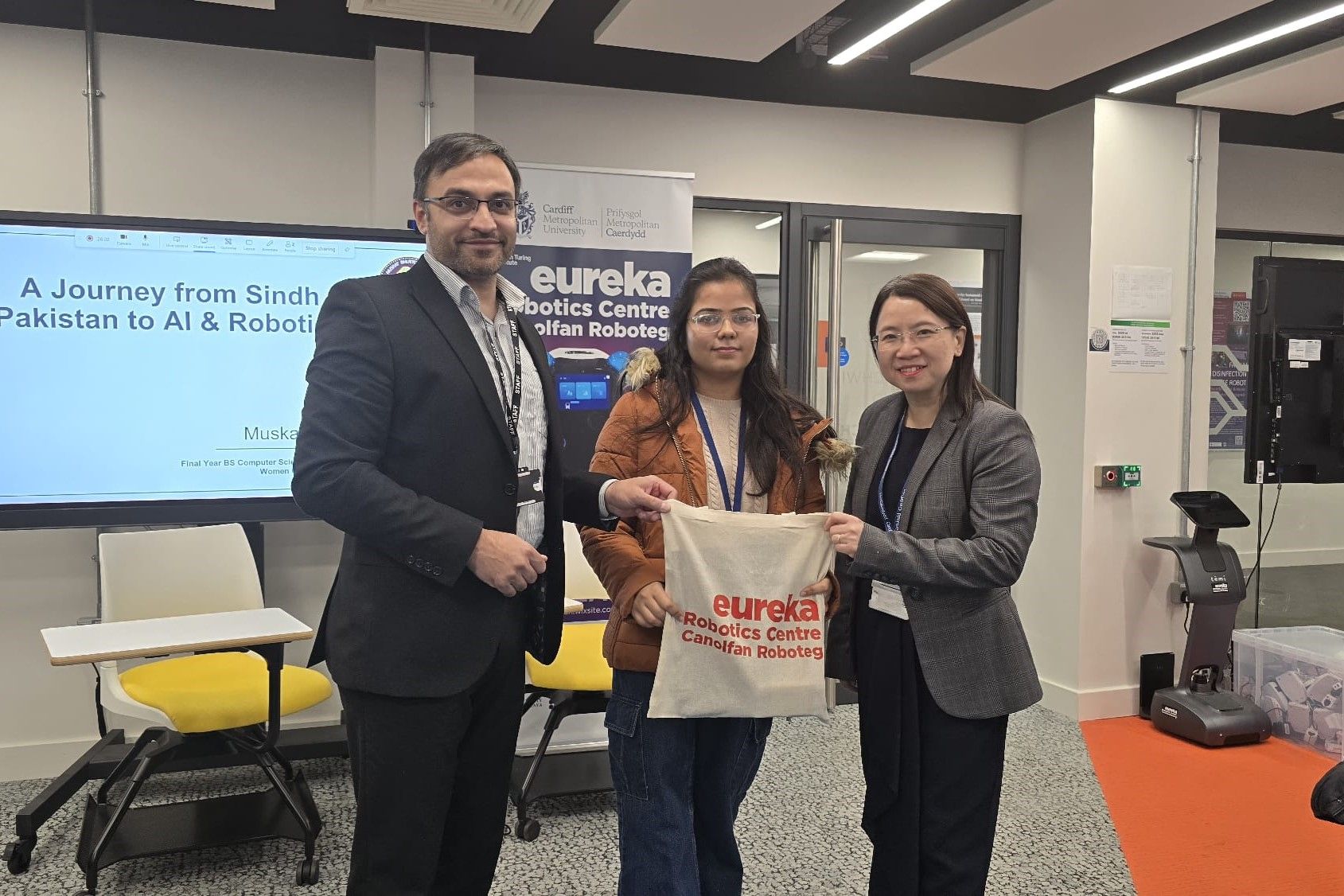 Three individuals stand for a photograph. The young woman in the centre holds up a branded tote bag