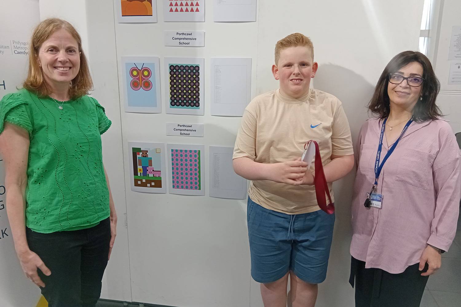 A young boy holding an award stands in front of a display of computer generated artwork. Standing on either side of him are staff from Cardiff Met.