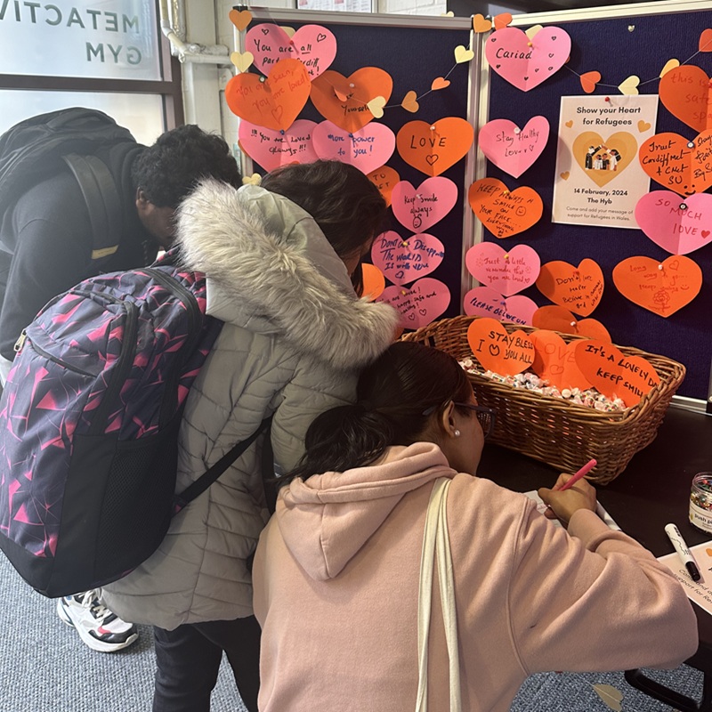 University students lean over a table and display to write messages of support for refugees on coloured, heart-shaped paper card