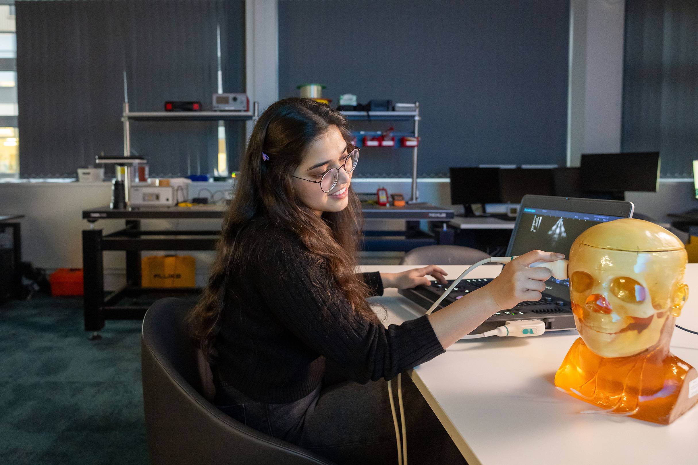 A student sat at a desk uses an ultrasound scanner on a simulated human skull. An open laptop computer in front of them shows the scan.
