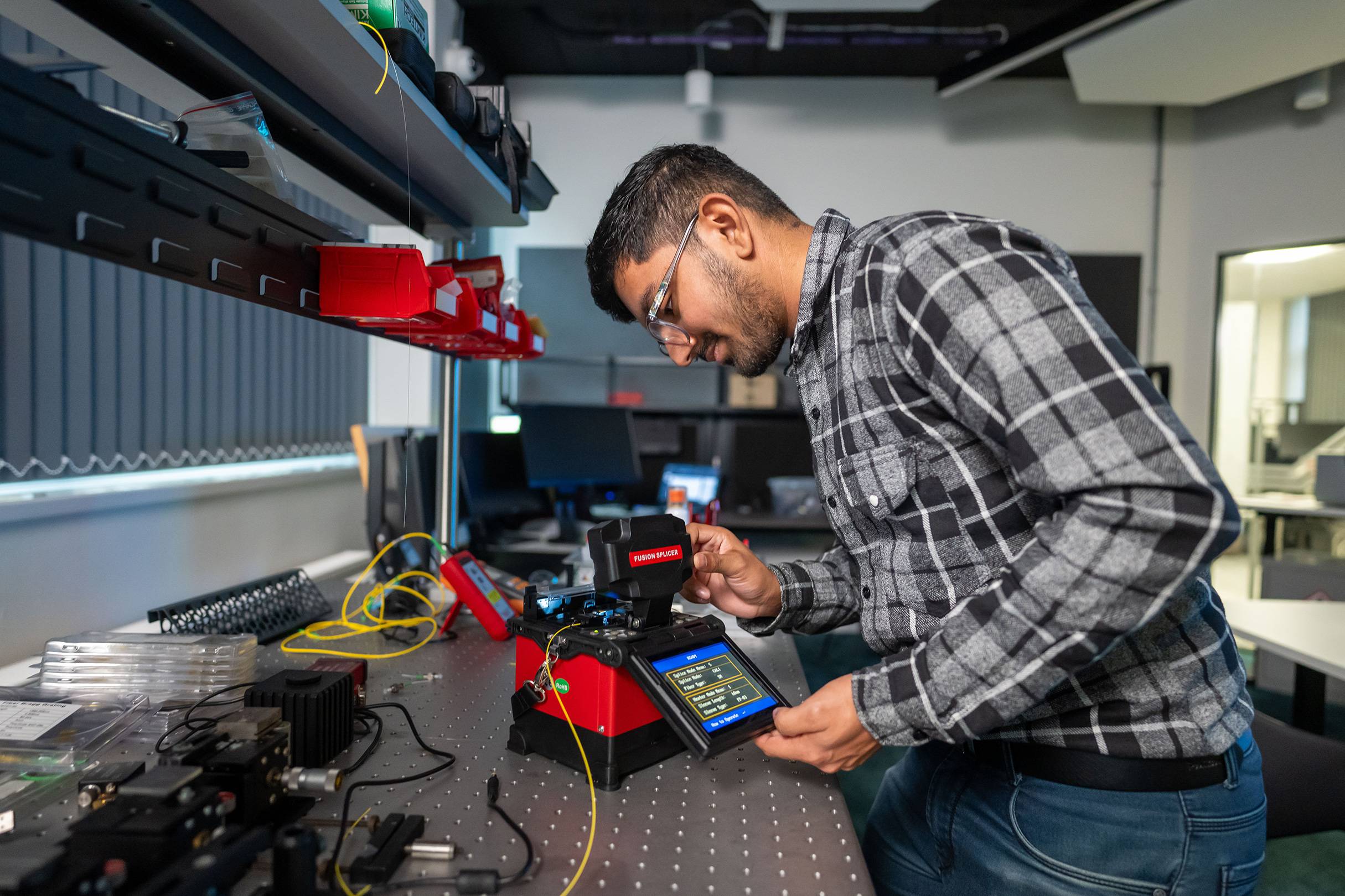 A student pressing the touchscreen of a small electronic device. In the background are other devices and computer monitors.