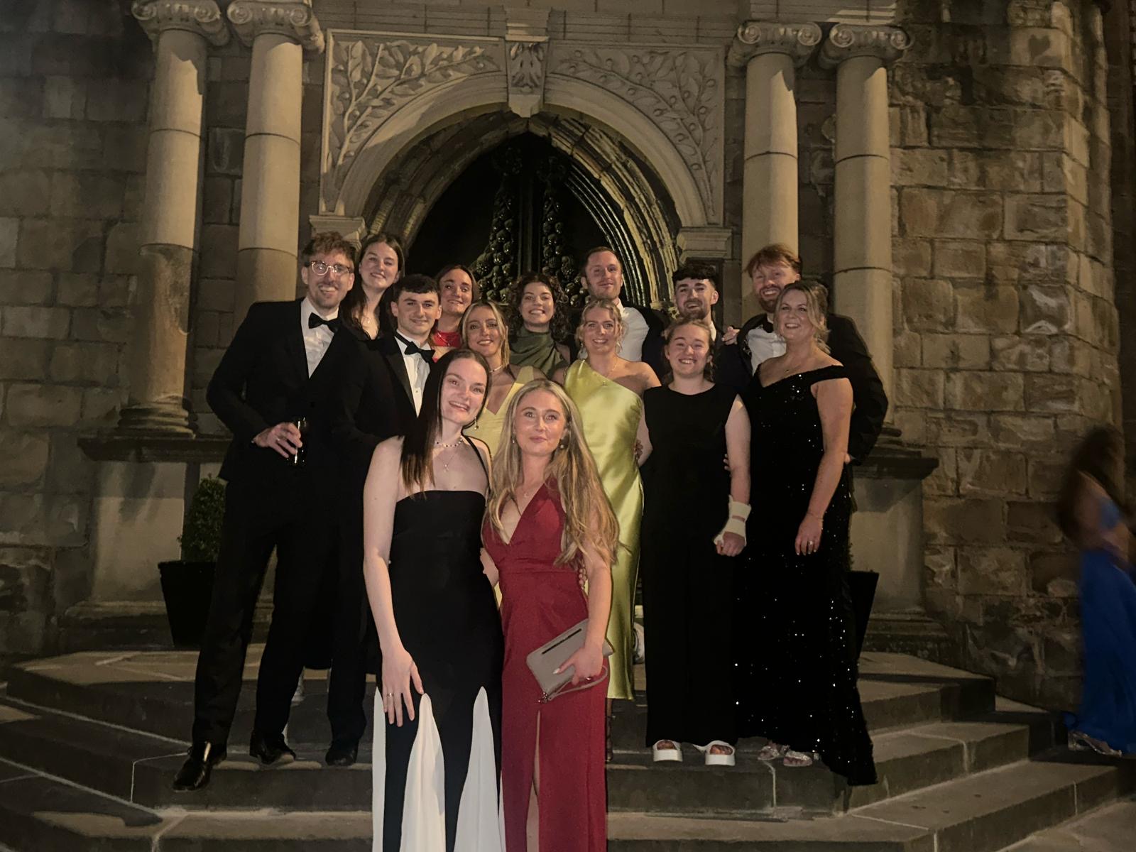 A group of Cardiff Met students and staff in formal suits and dresses stand on the steps in front of an ornate stone entrance