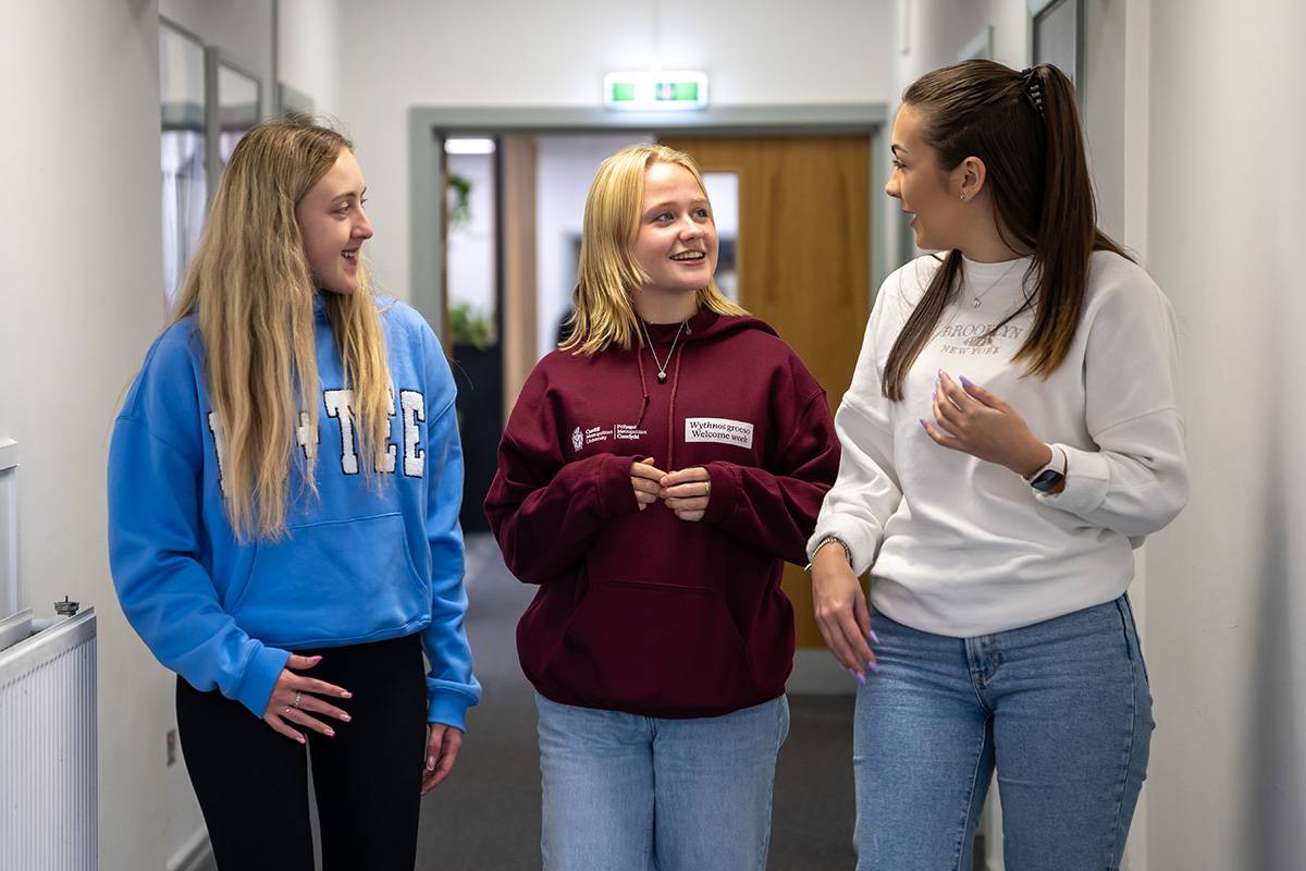 Three young women walk down a hallway together, wearing casual clothes and having a conversation.