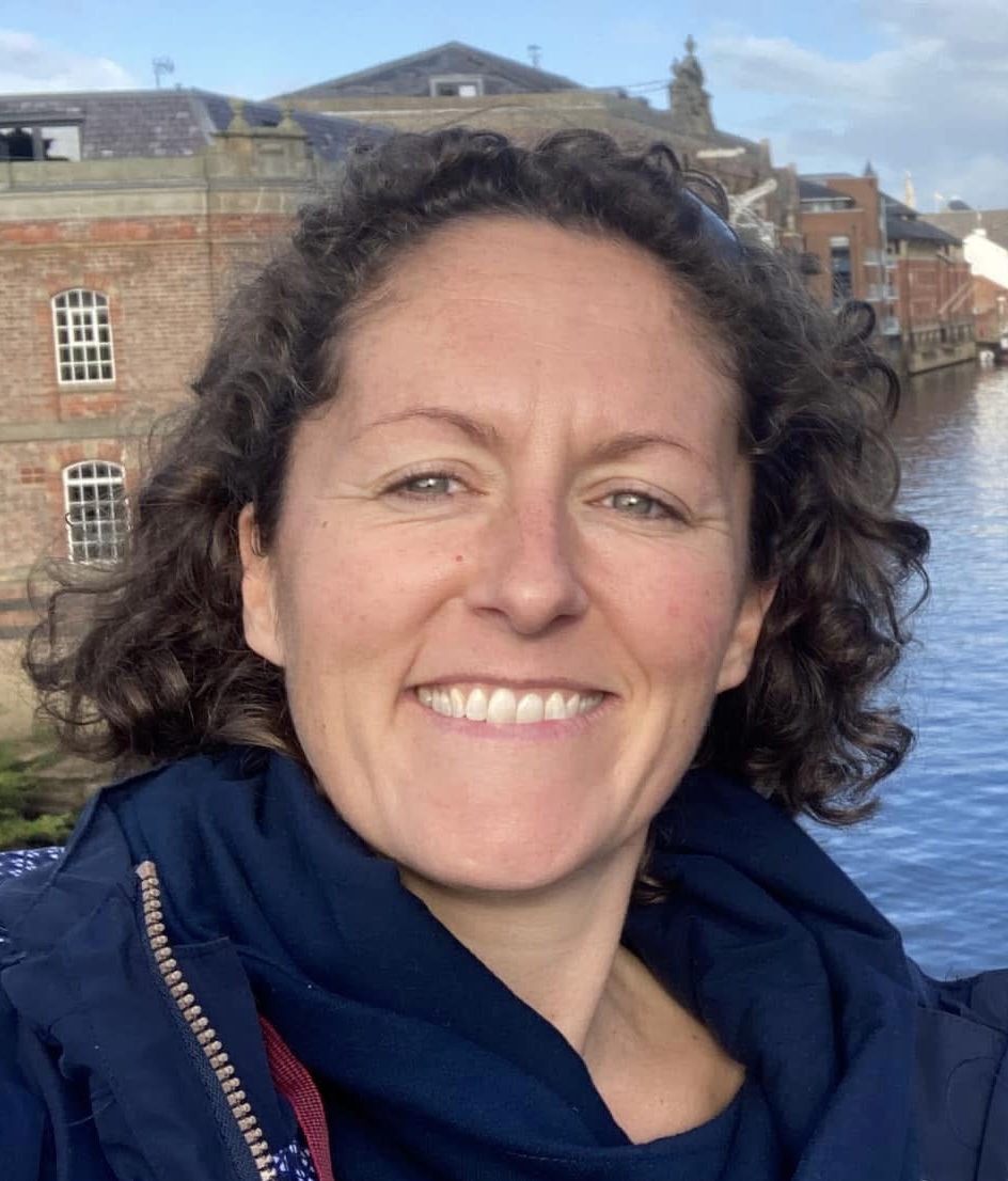 A woman with curly hair in a blue jacket stands by a river, with water flowing gently in the background.