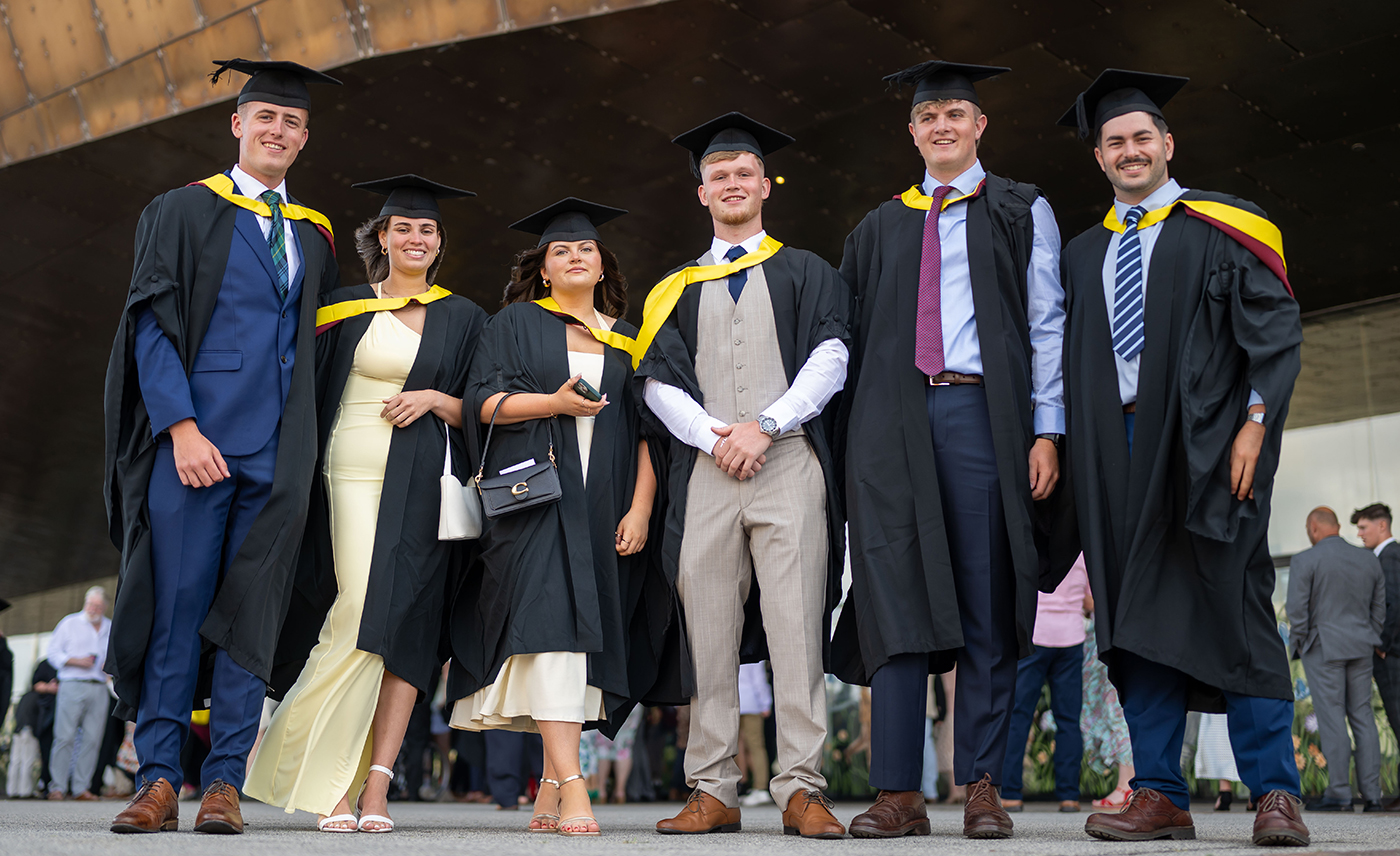 A group of six Cardiff Metropolitan University graduates in academic gowns and caps smiling and posing together outside the Wales Millennium Centre during the Class of 2025 graduation ceremonies.