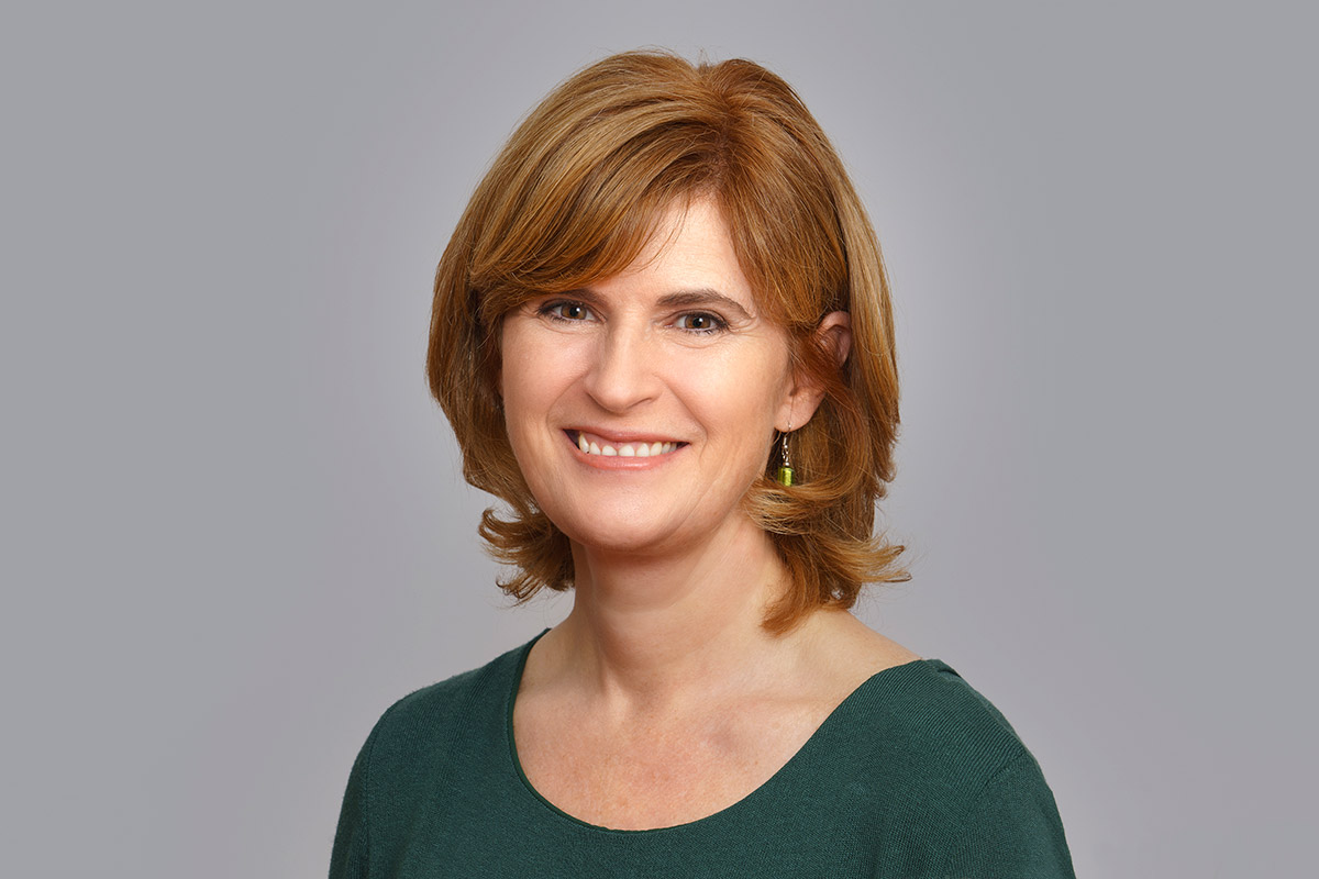 A woman with red hair wearing a green shirt smiles warmly at the camera.