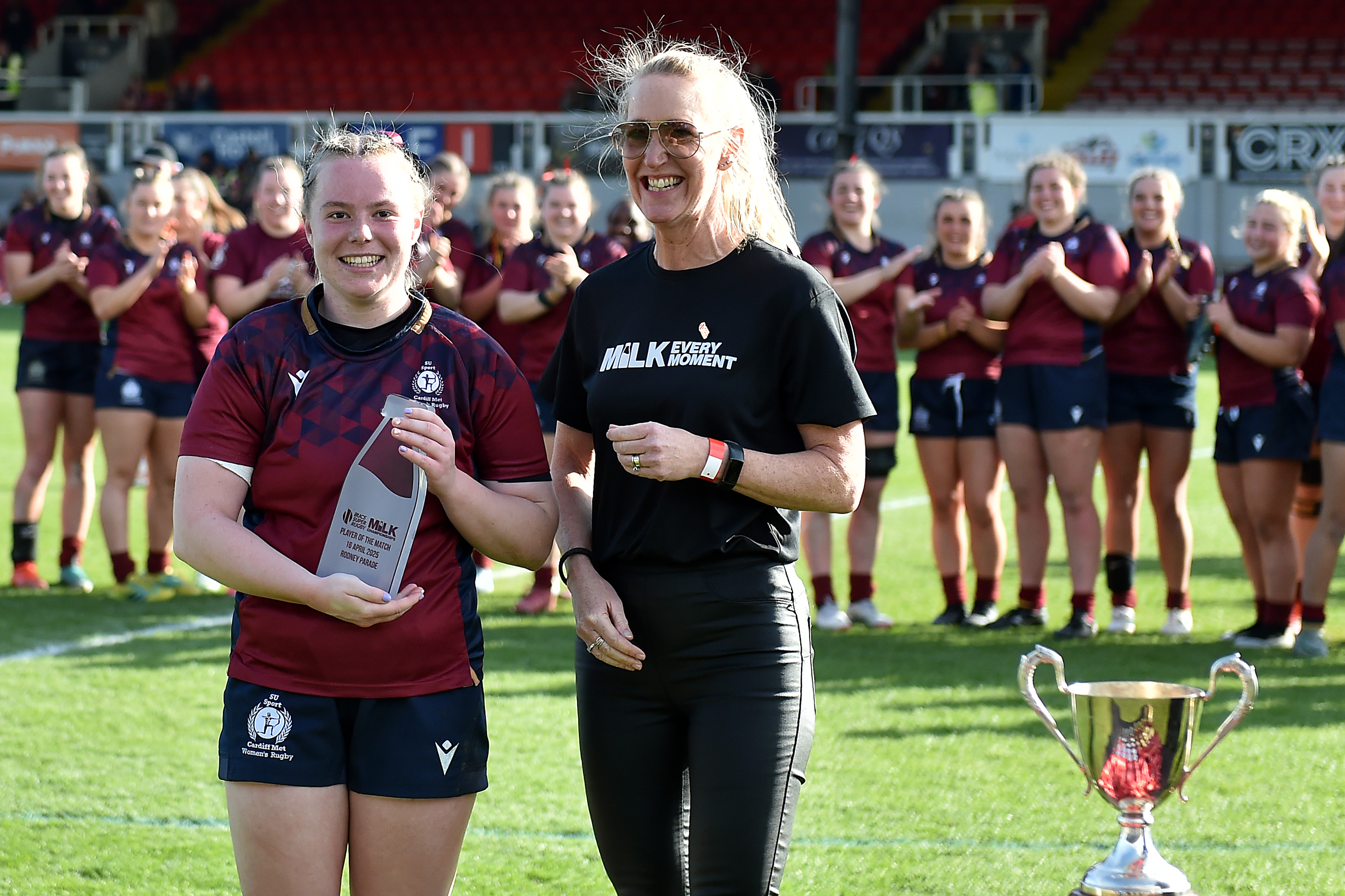 A female rugby player poses with an award in front of her teammates and alongside another individual
